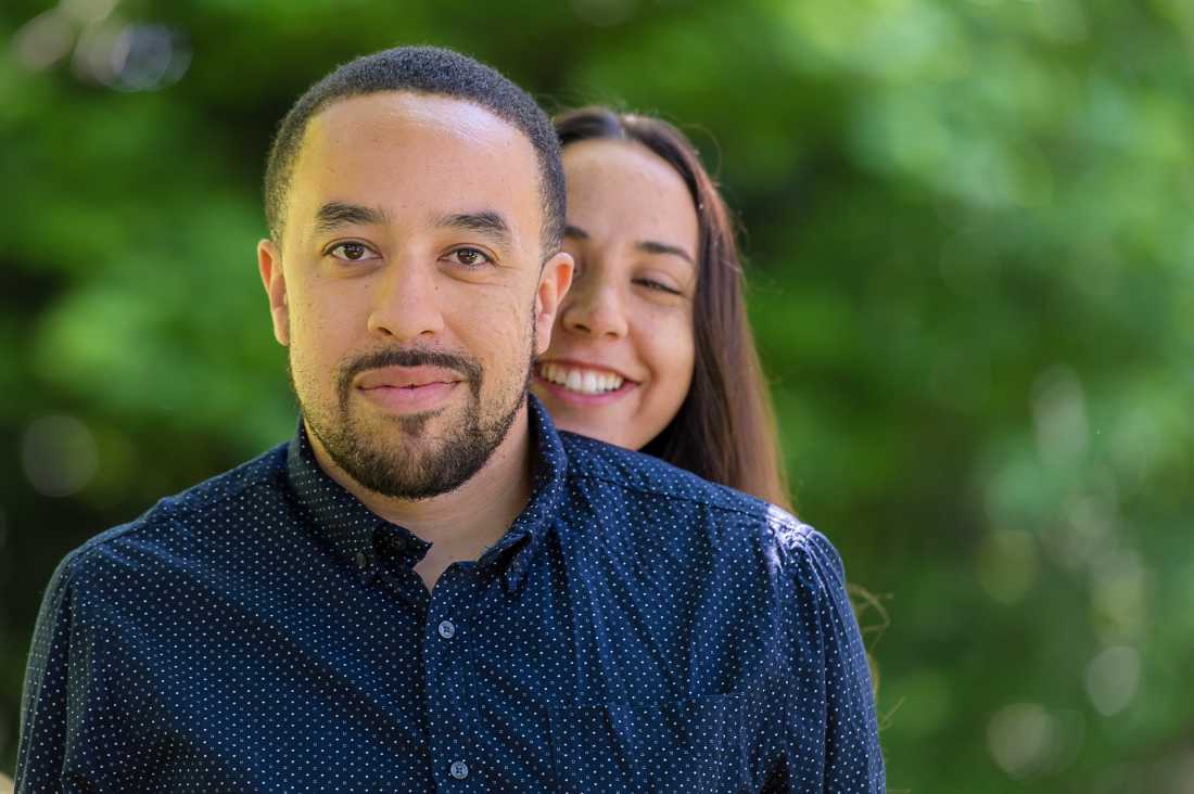 guy looking straight at the camera, girlfriend behind him tickling