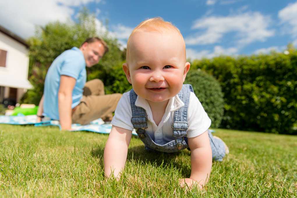 baby crawling towards camera across the grass, dad in background